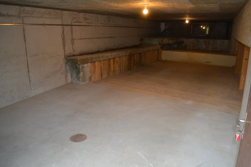 This image shows the interior of a garage or storage space. The space has a concrete floor and wooden walls, with a raised platform along one side. The lighting is dim, with a few overhead lights illuminating the space. There appears to be a drain or acce