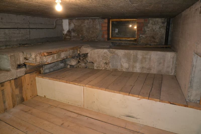 Unfinished basement with concrete walls, wooden floor, and a mirror on the wall. The space appears to be used for storage or utility purposes.