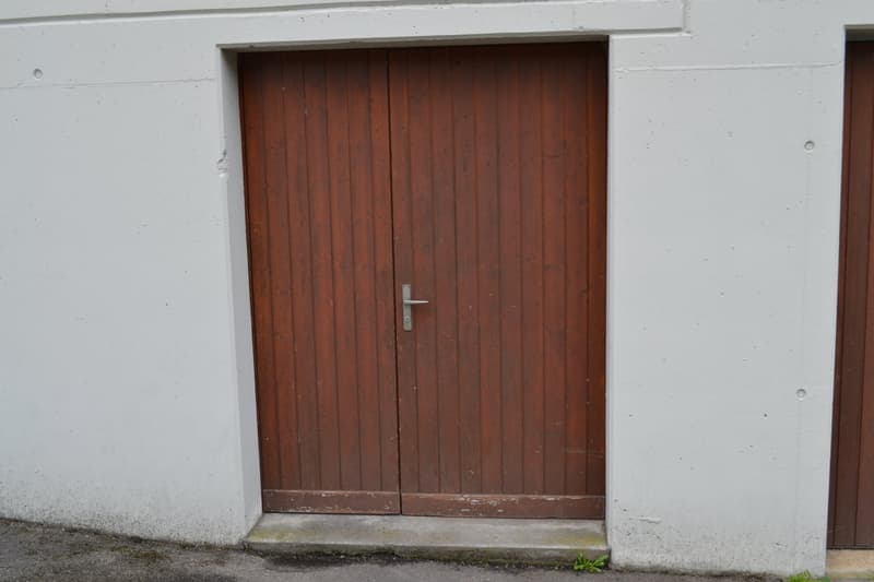 The image shows a wooden door set in a white wall. The door is made of vertical wooden planks and has a metal handle. There is a small step leading up to the door.