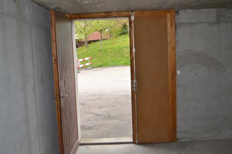 The image shows the interior of a garage or storage space. The space has a concrete floor and wooden walls, with a large open doorway leading to the outside. The view through the doorway shows a grassy area with some trees and a fence.