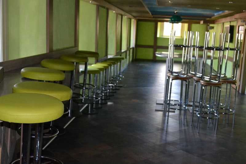 The image shows a long, narrow room with green walls and a dark hardwood floor. There are rows of yellow padded stools and chairs along the counter-style seating area, as well as several tall metal stools and tables. The room appears to be a restaurant or