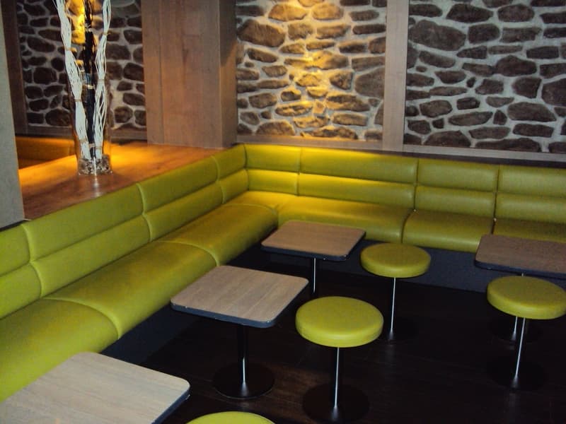 The image shows a cozy and modern interior space with a stone wall feature, a large green leather sectional sofa, and several small round tables with stools. The lighting is warm and creates a comfortable atmosphere. The visible features include a firepla