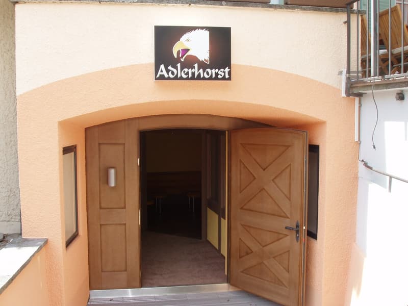 The image shows the entrance to a building called 'Adlerhorst'. The entrance has a large wooden double door with a decorative pattern. Above the entrance is a sign with the Adlerhorst logo, which features an eagle. The building has a beige exterior and ap