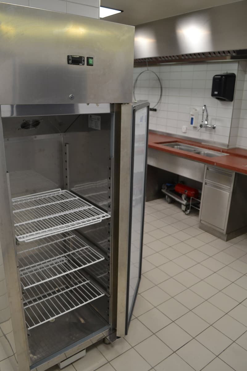 An empty stainless steel fridge in the middle of a kitchen with a sink and gas cylinder