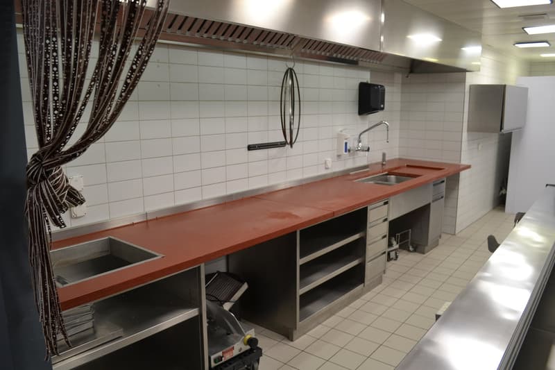Industrial kitchen with tiled walls, stainless steel countertops, sinks, ventilation hoods, built-in cabinets, countertop microwave, and various kitchen appliances.