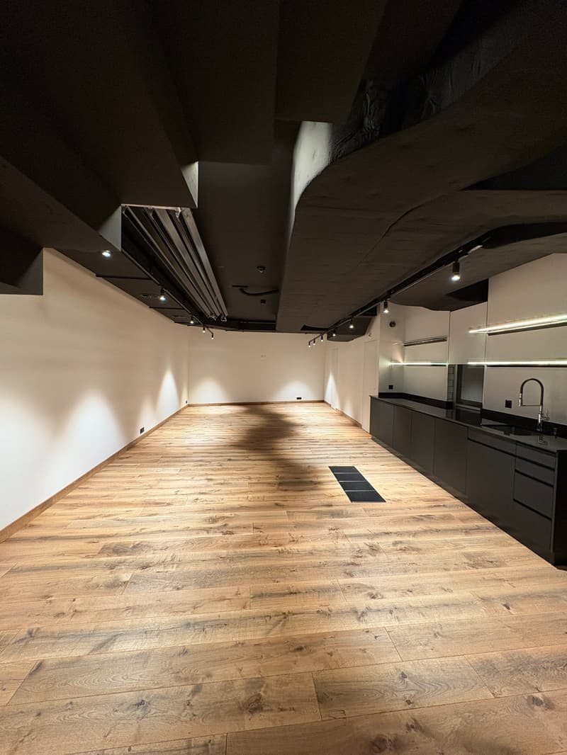 Empty room with hardwood floors, track lighting, black accent walls, open kitchen with dark cabinets and stainless steel backsplash, modern black matte sink with stainless steel faucet