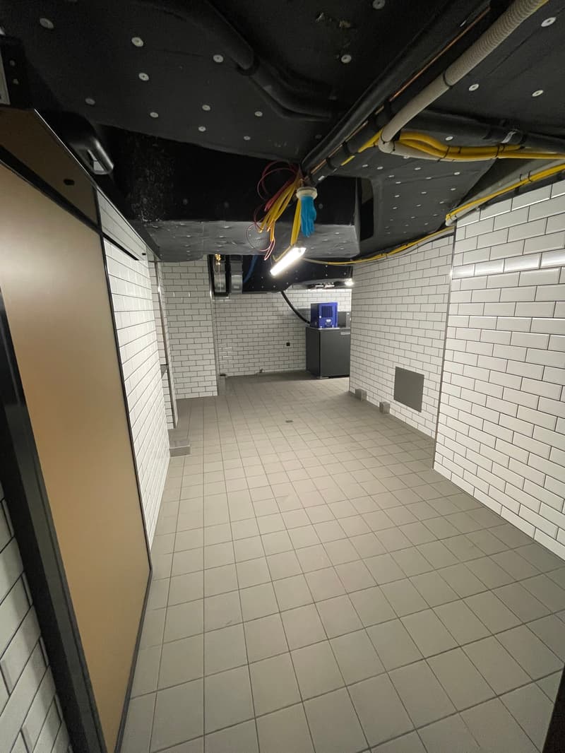 A vacant room with white tiled floors, white brick walls, a black ceiling with wires, a gray cabinet, and a bright white light on the ceiling.