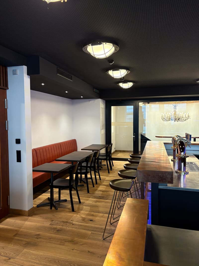 Dining area with red booth, black tables and chairs, wooden floor, ceiling lamps, bar with copper top and stools, entrance with glass doors, chandelier, white walls