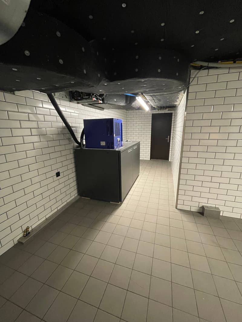 A room with white tiled walls and floor, an electronic device on a counter, and a door on the right.