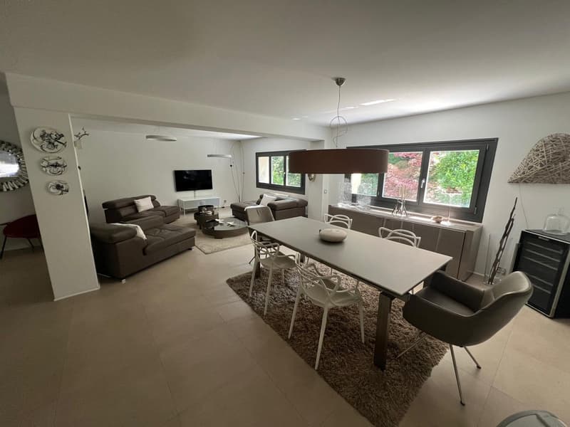 Open plan living and dining area with modern furniture, beige tiles, large windows, and a flat-screen TV.