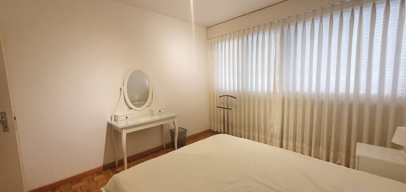 Bedroom with a bed, white desk with mirror, white nightstand, white curtains, wooden floor