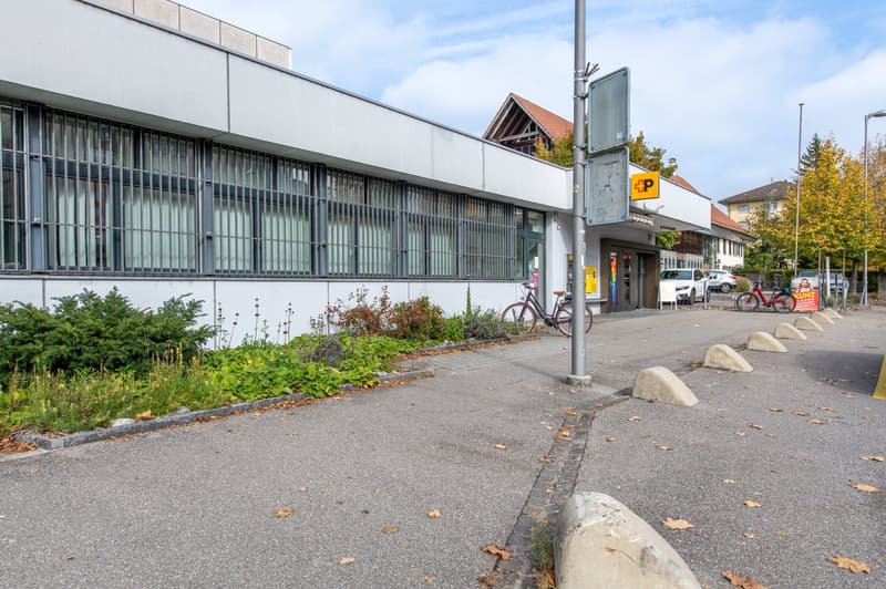 Building with many windows, bicycle parking in front, a few cars parked nearby, a sloped parking curbs, a pole with signages, fallen leaves on the ground