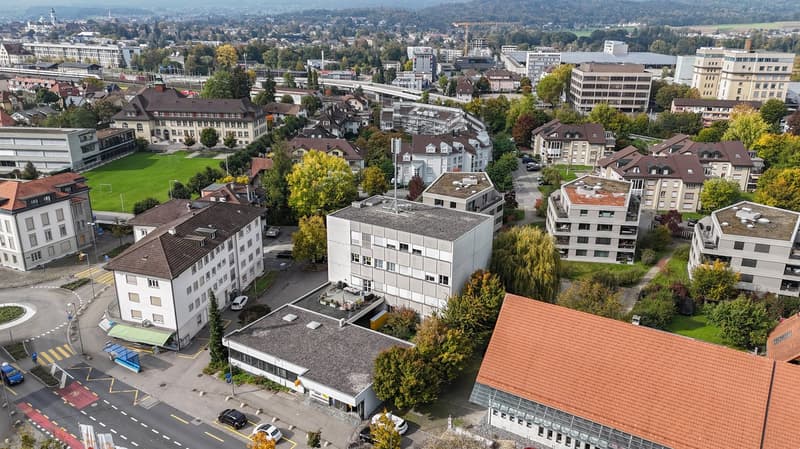 Urban city view, multiple residential buildings, roads, greenery, traffic, a crane visible in the distance, and mountains in the background.