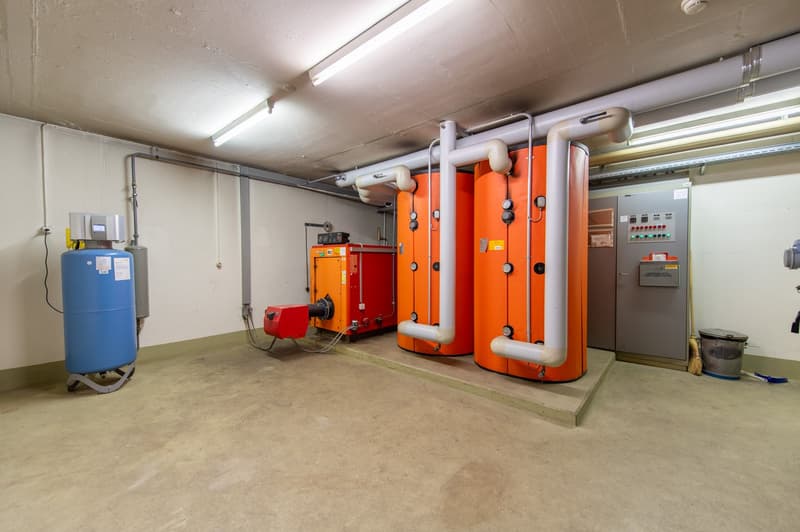 Large utility room with storage space, large heating installations, equipment and piping on the walls, cement floor