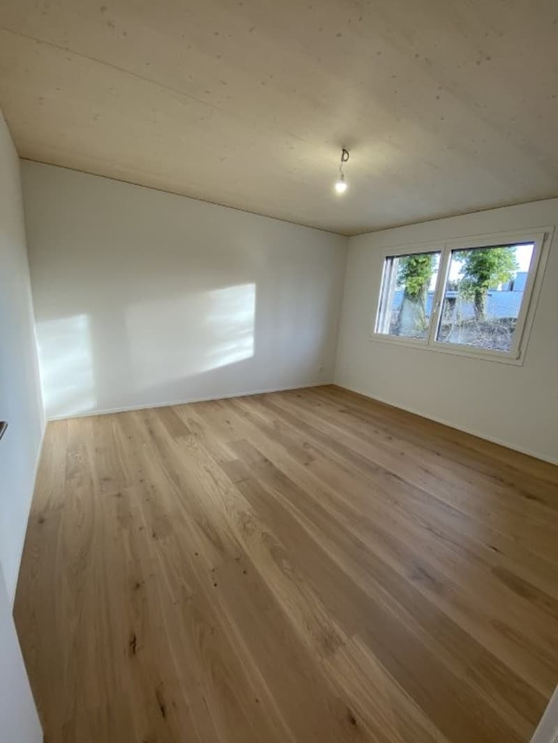 Spacious room with light-colored hardwood flooring, white walls, and a window providing natural light.