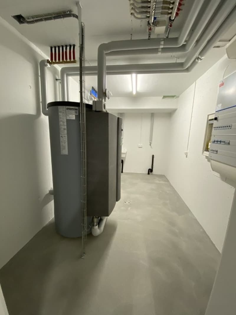 The image shows a utility room with a water heater, pipes, and electrical panels visible. The room appears to be a functional space for housing building utilities and equipment.