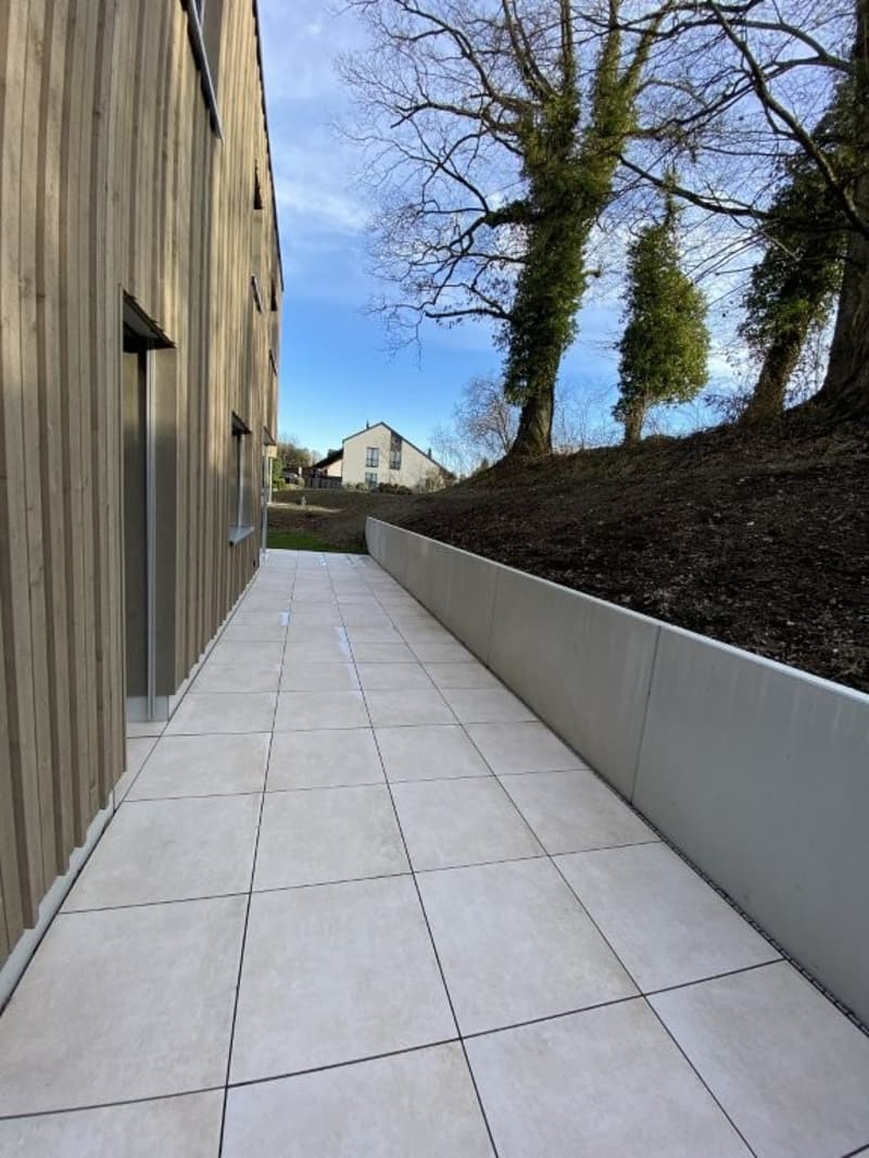 The image shows a paved walkway made of concrete tiles. The walkway is surrounded by a raised concrete wall on one side, and there are trees and a building visible in the background.