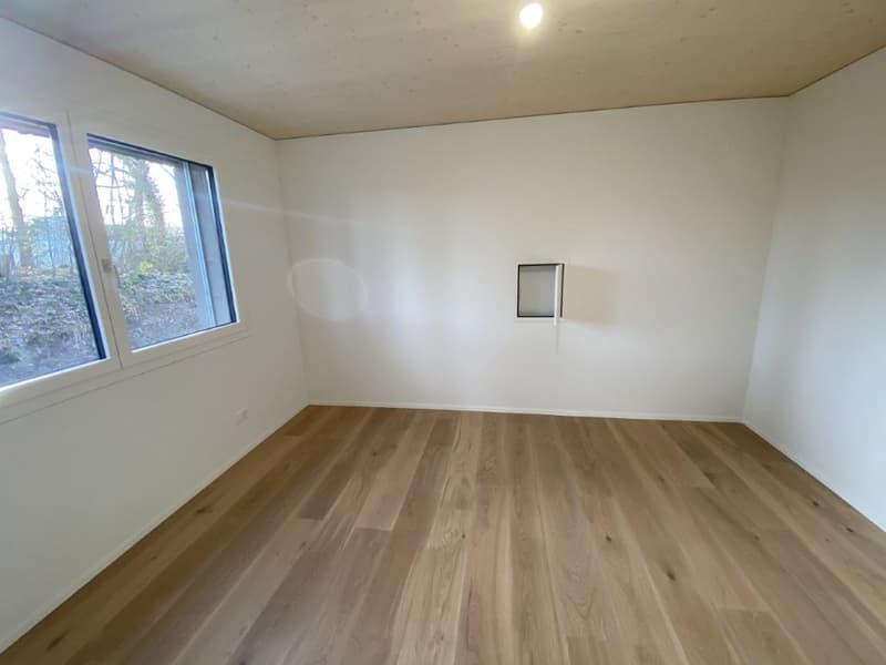 This image shows a spacious room with white walls and a hardwood floor. The room has a large window that provides natural light and a view of the outdoors. The room appears to be empty, with no furniture or decor visible.