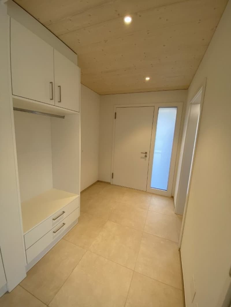 The image shows a hallway with white walls, a wooden ceiling, and a tiled floor. There is a built-in closet or storage unit along one wall, and a door leading to another room. The hallway appears to be well-lit with recessed lighting.