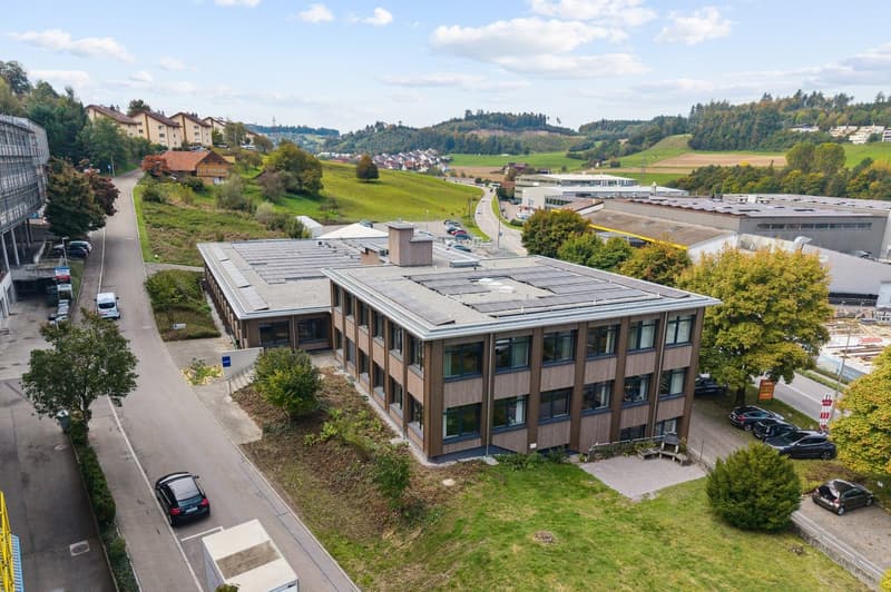 2 story office building, solar panels on the roof, surrounded by trees and bushes, parking lot, road, several cars parked