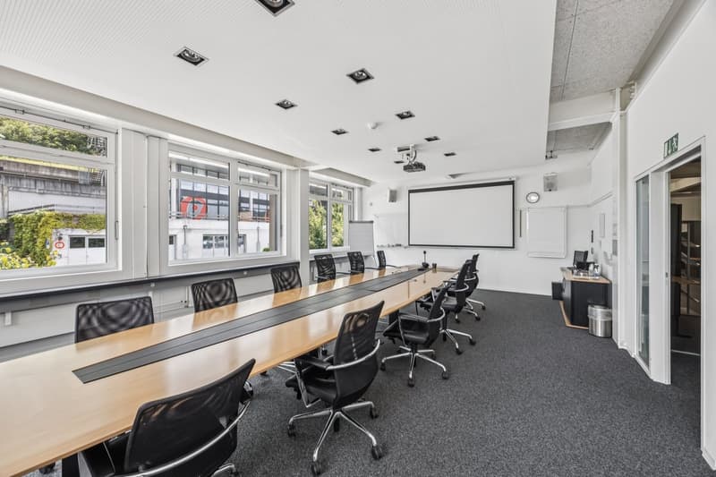 Large conference room with a long wooden table, black office chairs, and a projector screen on the wall. The room has large windows and recessed lighting fixtures on the ceiling.