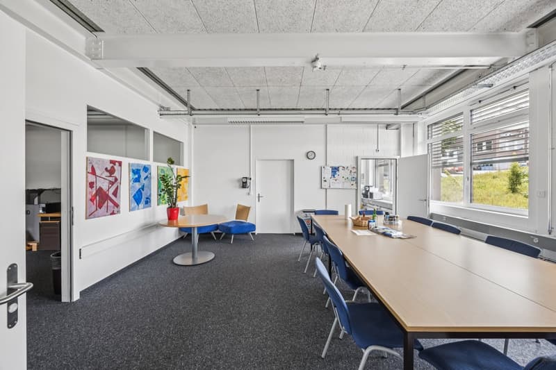 Large open office space with white walls, gray ceiling tiles, and carpeted floor. The room has a conference table with blue chairs, a round table with chairs, and artwork on the walls. There are windows providing natural light and various office equipment