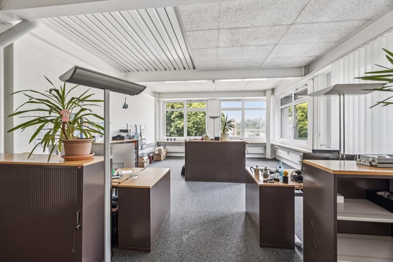 Spacious office space with large windows, modern furniture, and kitchen appliances including a dishwasher. The room has a bright and open layout with a mix of work surfaces and storage areas.