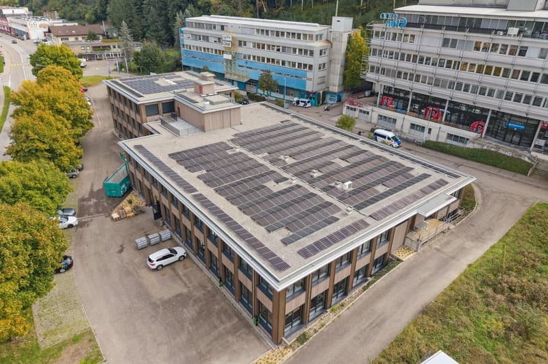 Building with solar panels, several cars parked, green sign, trees and grass, street in background
