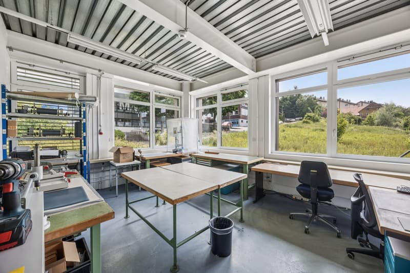 Large open space with high ceilings, concrete floor, and large windows providing a view of the surrounding greenery. The room is equipped with various workbenches, shelves, and tools, suggesting it is used as a workshop or studio space.