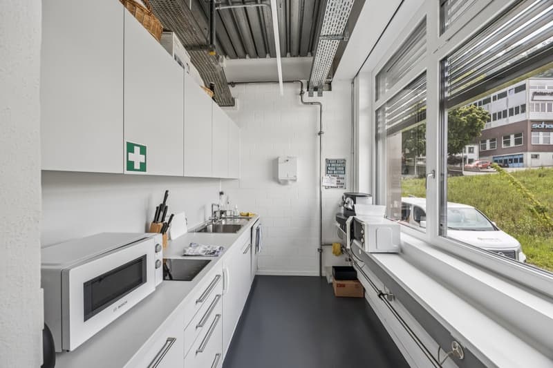 Compact kitchen with white cabinets, stainless steel appliances including a dishwasher, sink, and microwave. The kitchen has a long narrow layout with a window overlooking the street outside.