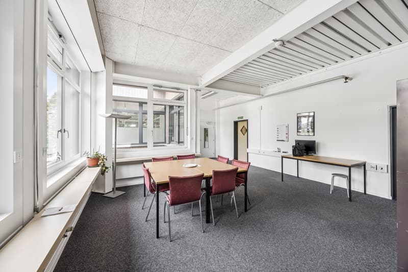 Large open space with a conference table and chairs, white walls, gray carpeted floor, and large windows providing natural light. The room also has a TV mounted on the wall.