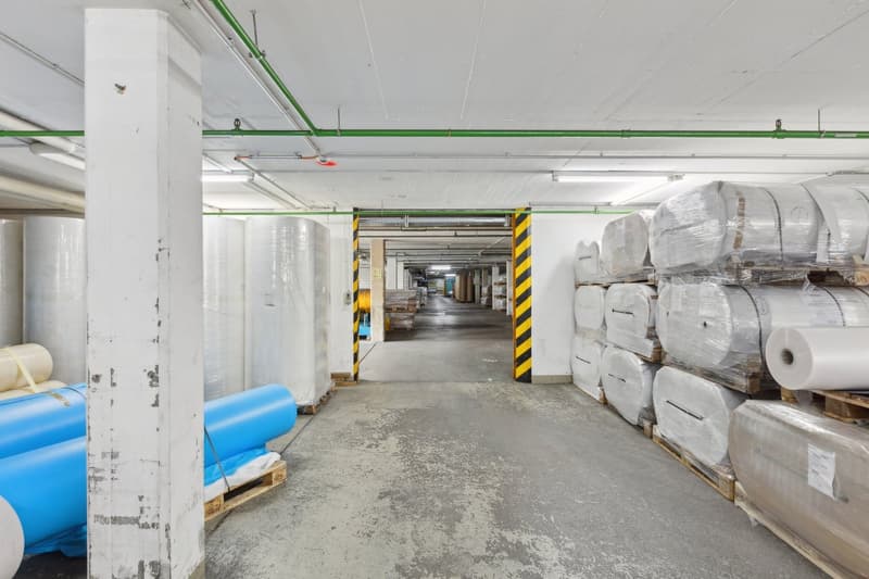 Large industrial warehouse space with concrete floors, white walls, and overhead pipes and lighting. The space contains various storage items and materials, including rolls of material and other supplies. There is a visible garage door entrance leading to