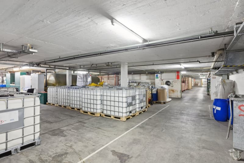 Large industrial warehouse space with concrete floor, overhead lighting, and various storage containers and equipment on pallets.