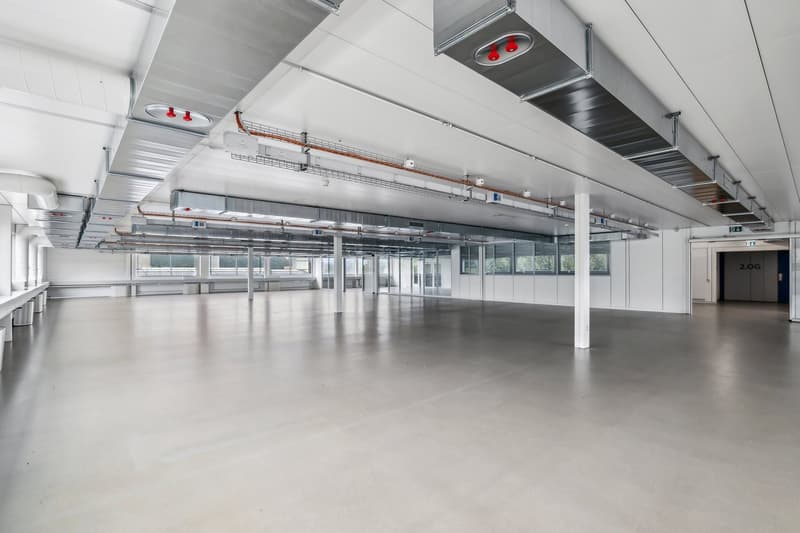 Large open commercial space with high ceilings, exposed ductwork, and concrete flooring. The space appears to be a warehouse or industrial facility with multiple overhead lights and windows along the walls.