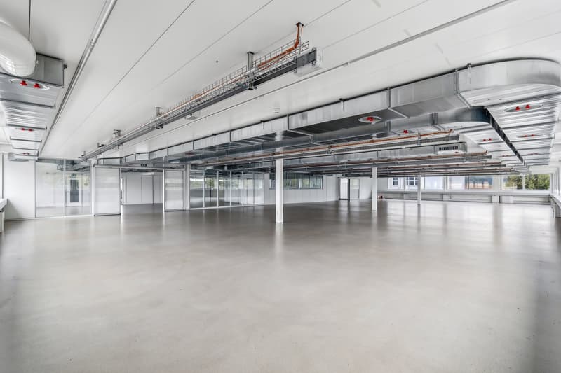 Large open commercial space with high ceilings, exposed ductwork, and concrete flooring. The space appears to be a warehouse or industrial facility, with multiple overhead lighting fixtures and a clean, minimalist design.
