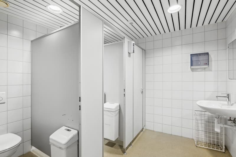 The image shows a bathroom with white tiled walls and a slatted ceiling. It has a toilet, a sink, and a paper towel dispenser on the wall. There are also some storage cabinets and a wire basket visible.