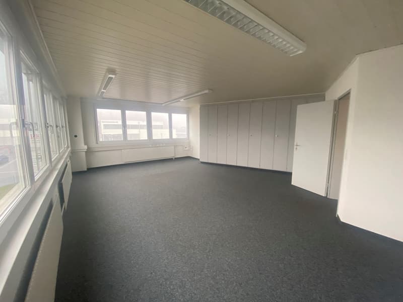 Empty room with white walls and ceiling, radiators along the walls, overhead lighting, large windows on one side