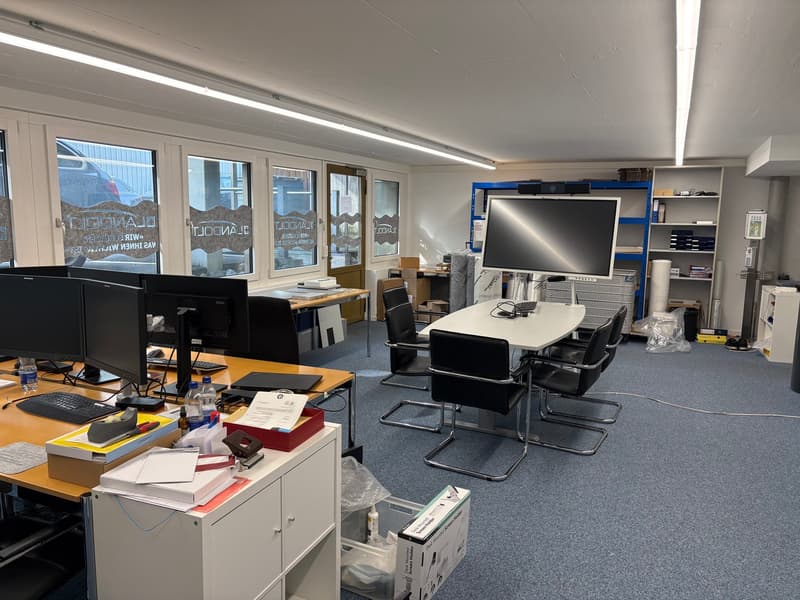 Office with blue carpet, white desk, black chairs, computer monitors, printer, shelves, boxes, and office supplies.