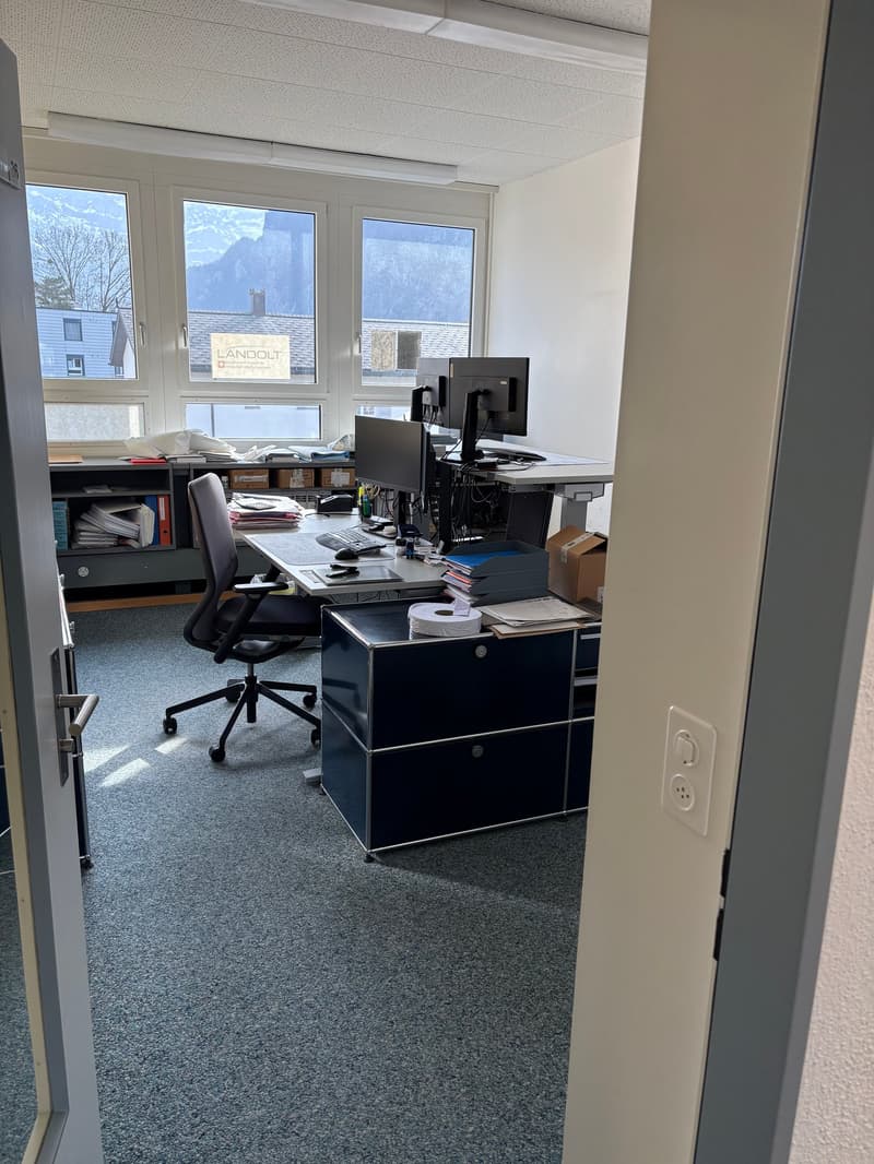 An empty office with a desk, monitor, keyboard, mouse, chair, papers, books, and a cabinet with drawers.