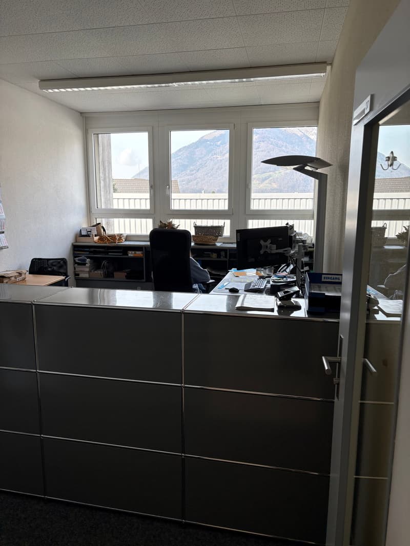 A spacious office with a desk, chair, computer monitor, books, and a lamp. There are two glass windows with a view of the mountains.