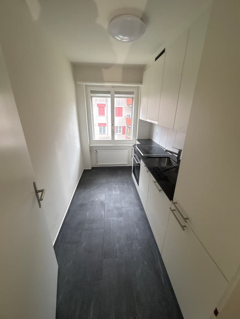 An empty kitchen with dark tiled flooring, white cabinets, black countertops, a window, a stove, an oven, a faucet, and a sink.