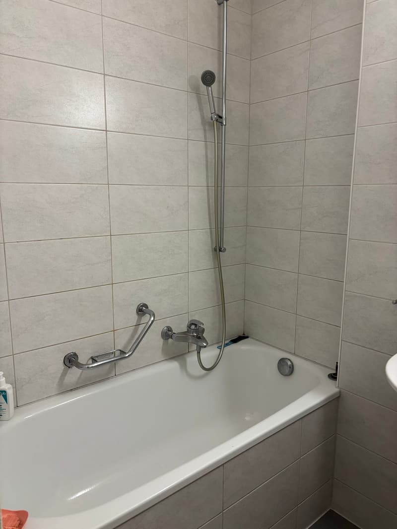 modern bathroom with white tiles, bathtub with showerhead, silver faucets, grab bars, toothbrush, soap