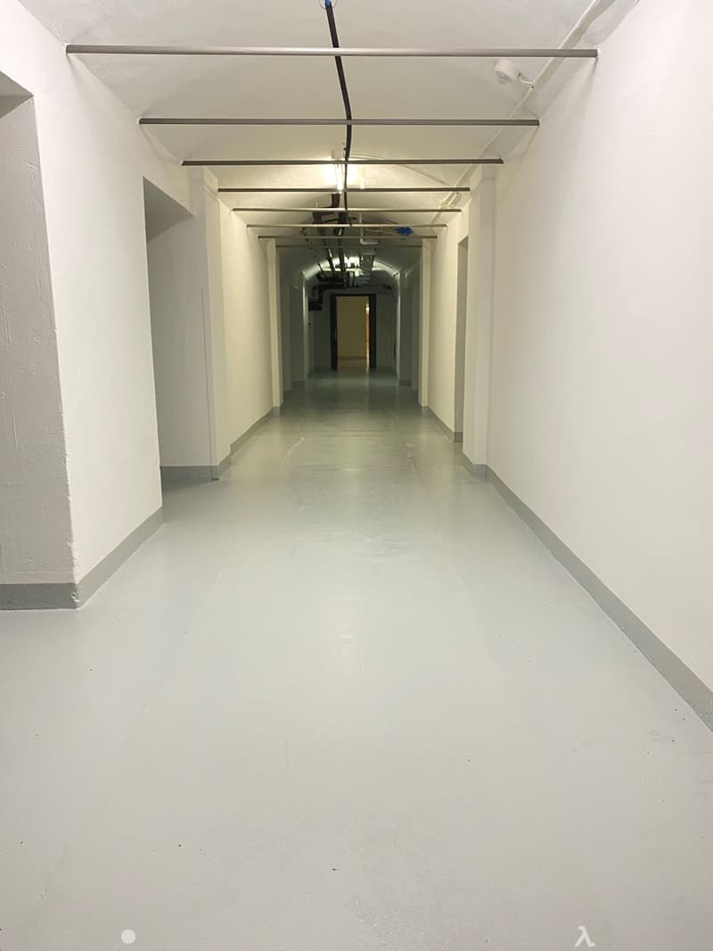 The image shows a long, narrow hallway with white walls and a concrete floor. The hallway has a series of recessed lighting fixtures in the ceiling, creating a well-lit and clean-looking space.