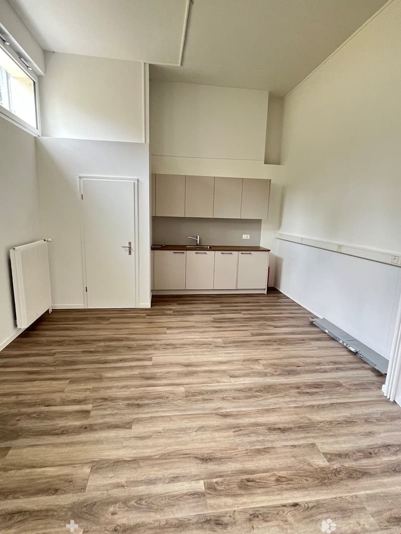 The image shows a studio apartment with a kitchenette area, hardwood-style flooring, and a white interior. The kitchenette features built-in cabinets and a sink. There is a door leading to another room or area. The overall space appears clean and minimali