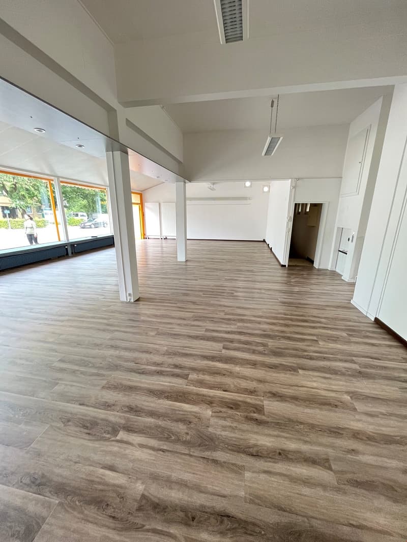 Large open space with hardwood-style flooring, white walls, and recessed lighting. The space appears to be a commercial or retail property with a large window overlooking the street.