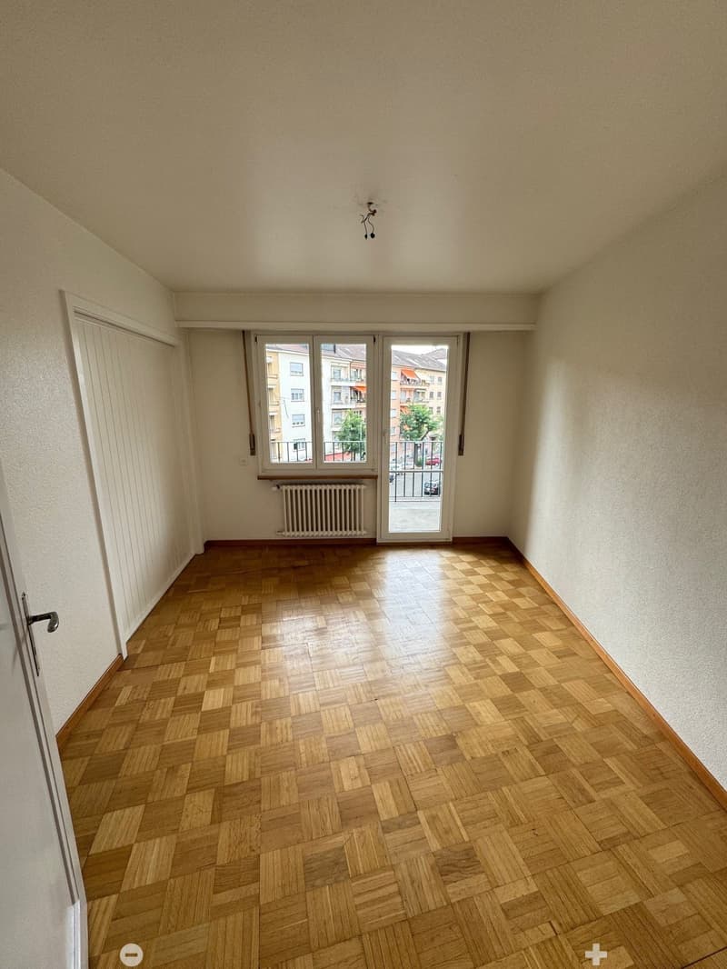 This image shows an empty room with a hardwood parquet floor, a sliding glass door leading to a balcony or terrace, and a radiator on the wall. The room has plain white walls and a simple light fixture on the ceiling.