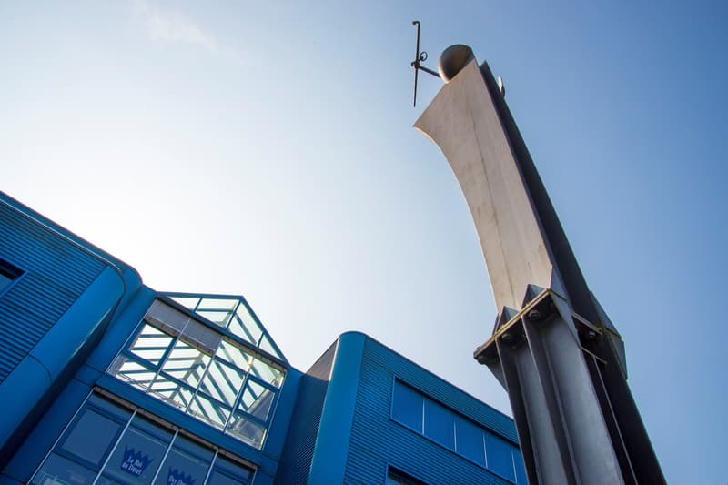A modern, multi-story building with a distinctive blue and glass facade. The building features a large, angular sculpture or architectural element on the roof, adding to its unique design.