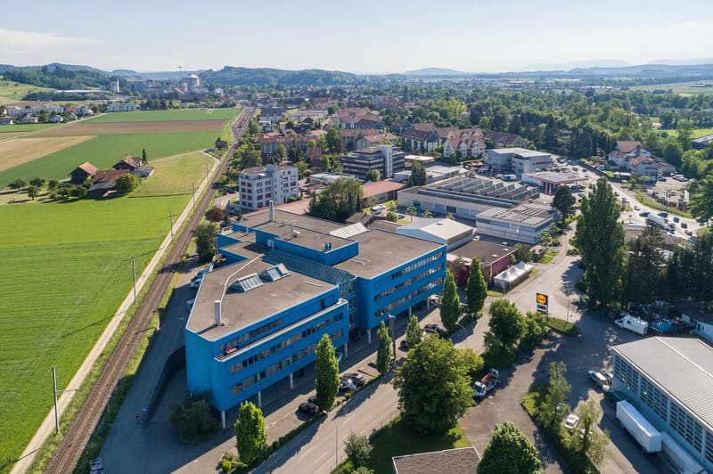 Aerial view of a city with a mix of residential and commercial buildings, surrounded by green fields and hills. The image shows a large blue commercial building with a parking lot in the foreground, and the city center with various buildings and infrastru