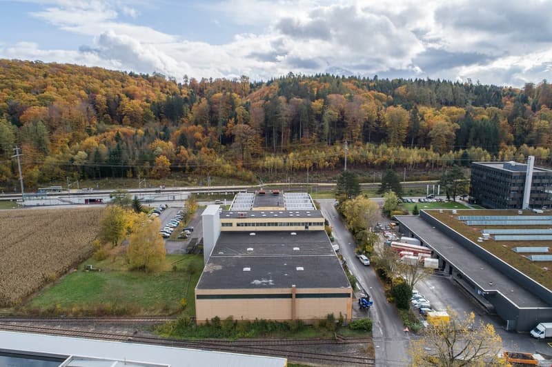 A large industrial building with green roofs, a spacious parking lot, surrounded by an extensive green field and forested hills in the background.