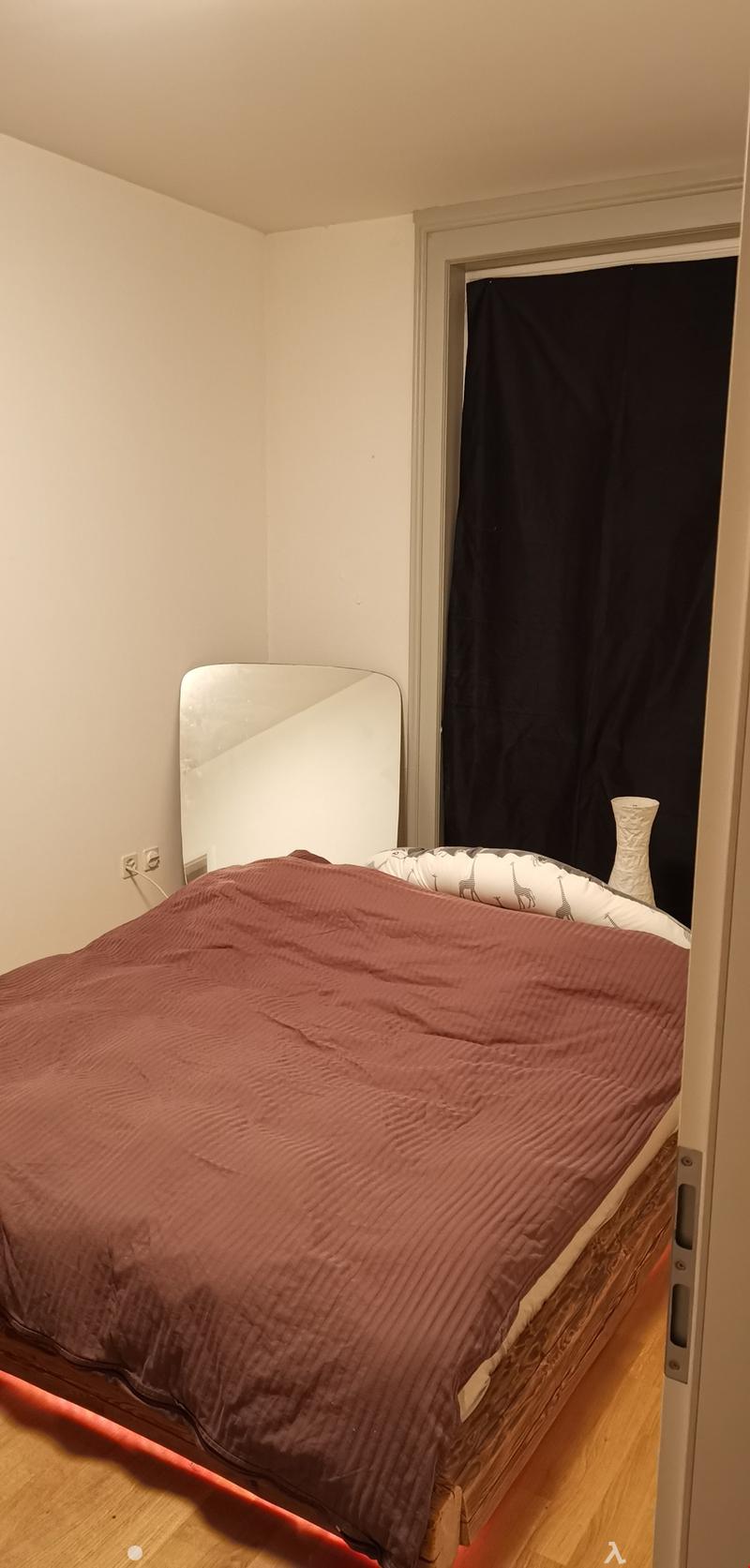 Bed with brown sheet, wooden floor, wall mounted mirror, white wall, curtain and a white vase